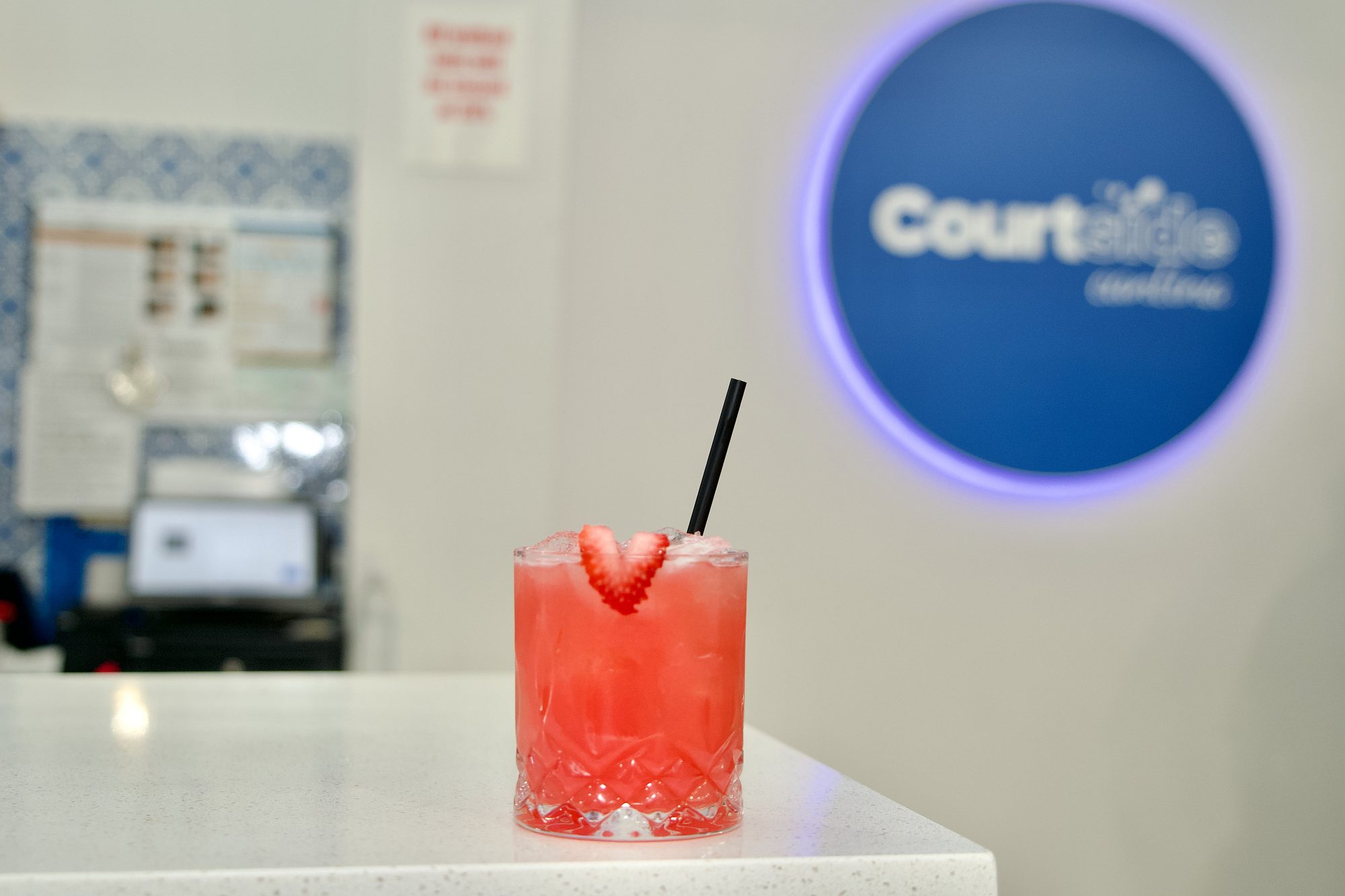 A red cocktail on the bar in front of the glowing Courtside Cantina sign